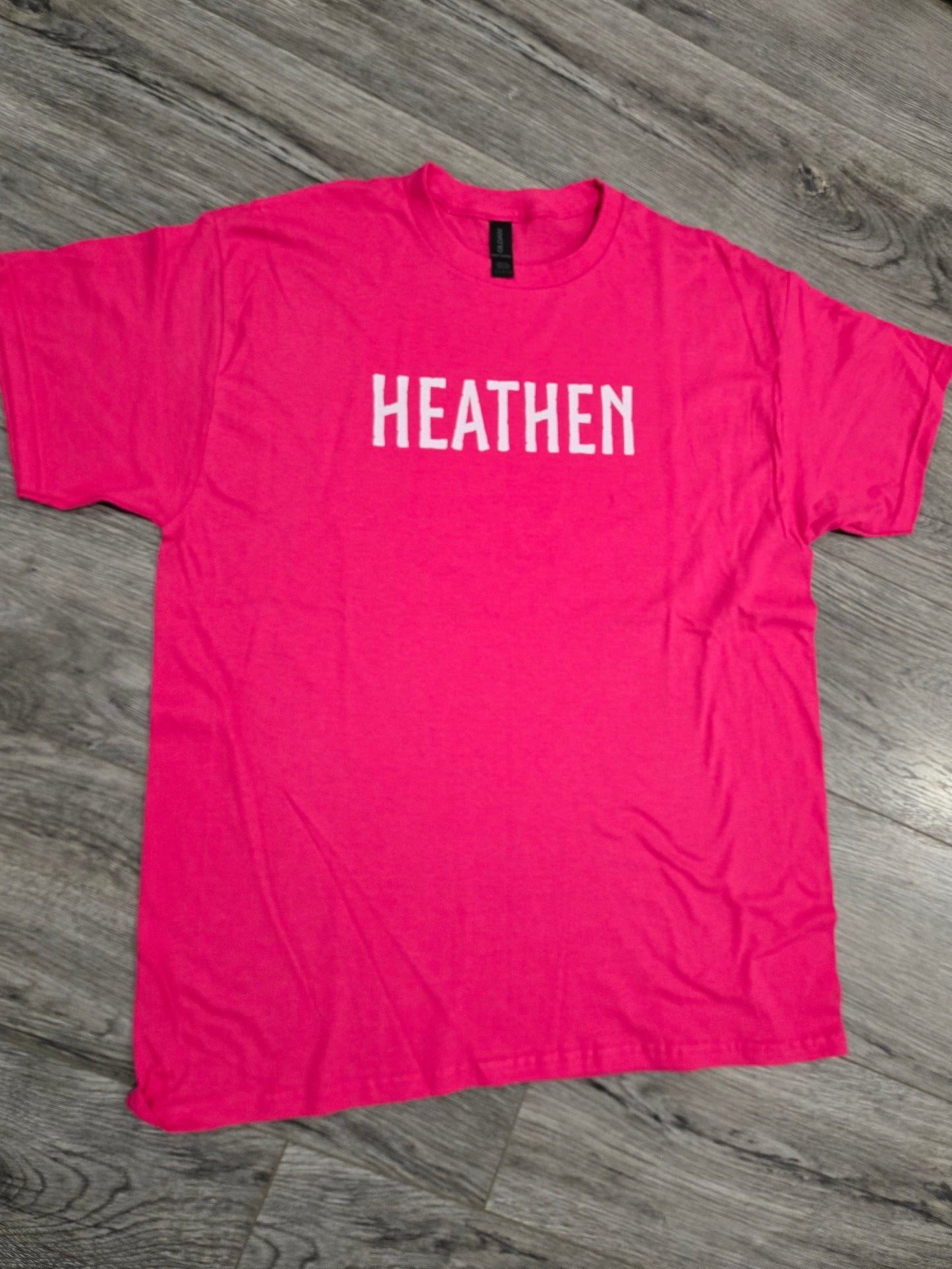 Pink t-shirt with 'HEATHEN' printed on it laid flat on a wooden floor.