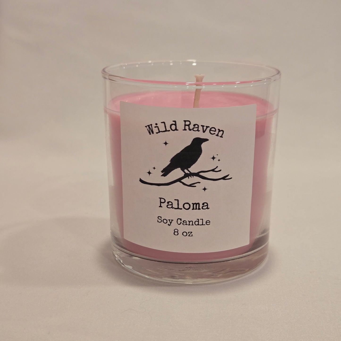 A pink soy candle in a clear glass jar with a white label that reads 'Wild Raven Paloma Soy Candle 8 oz' and features a black raven silhouette, photographed on a plain light background.