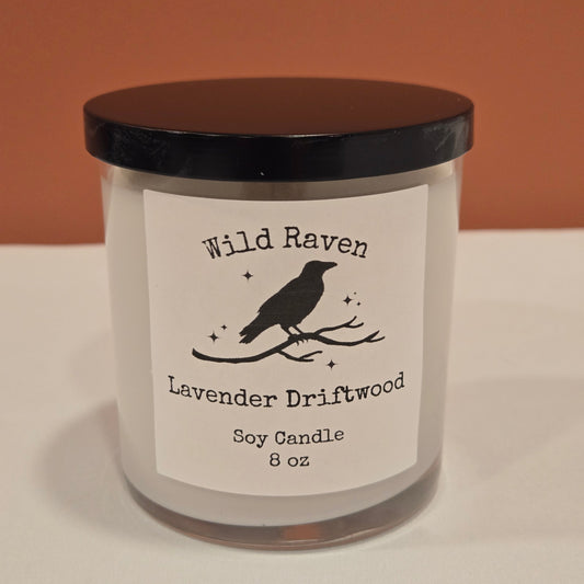 An 8 oz soy candle in a clear glass jar with a black lid and a white label featuring a black raven silhouette. The label reads 'Wild Raven' and 'Lavender Driftwood'.
