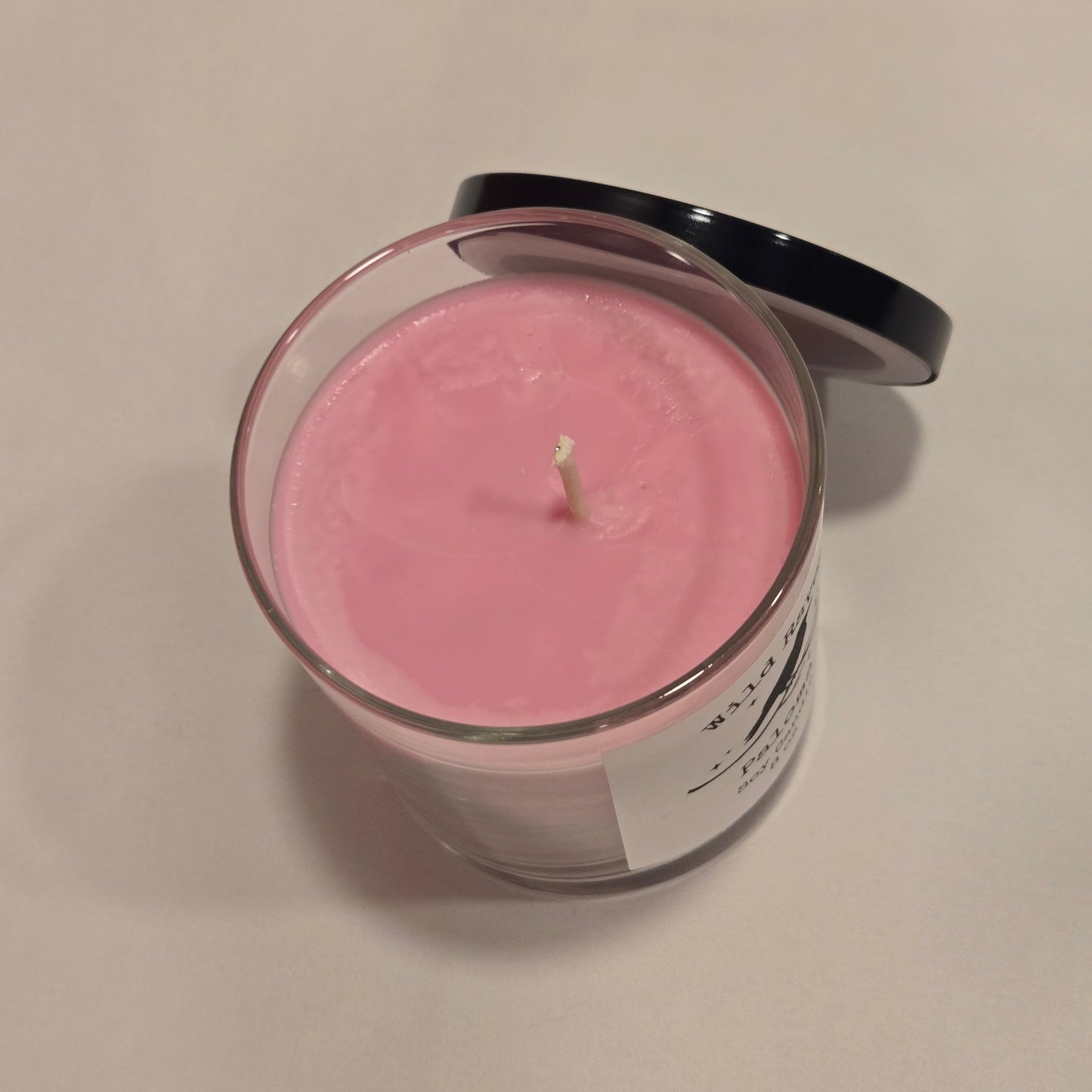 A pink wax candle in a clear glass jar with a white label and a black lid, photographed on a plain light background.