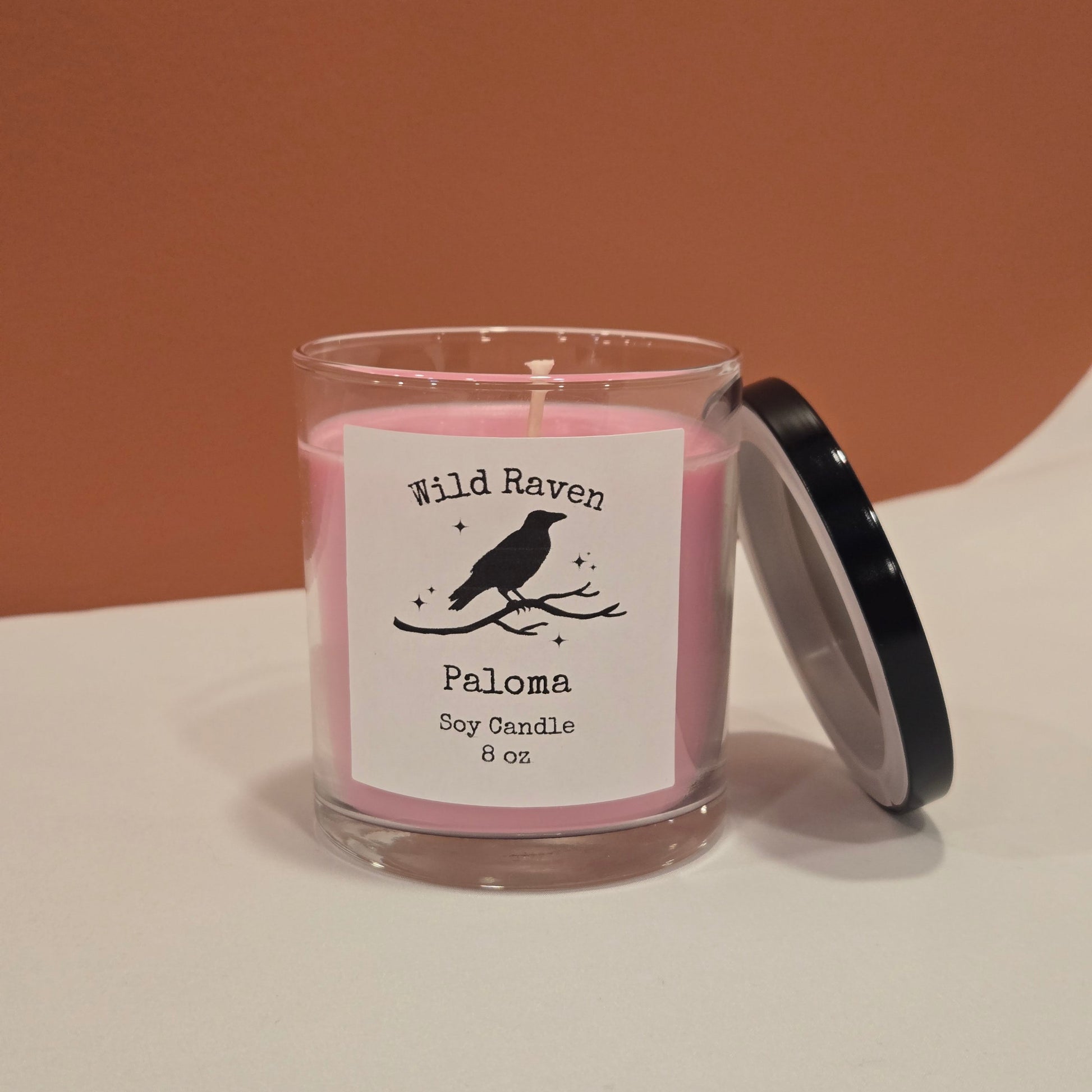 A pink soy candle in a clear glass jar with a black lid leaning against it, shown on a two-tone beige and terracotta background. The label reads 'Wild Raven' and 'Paloma Soy Candle 8 oz'.