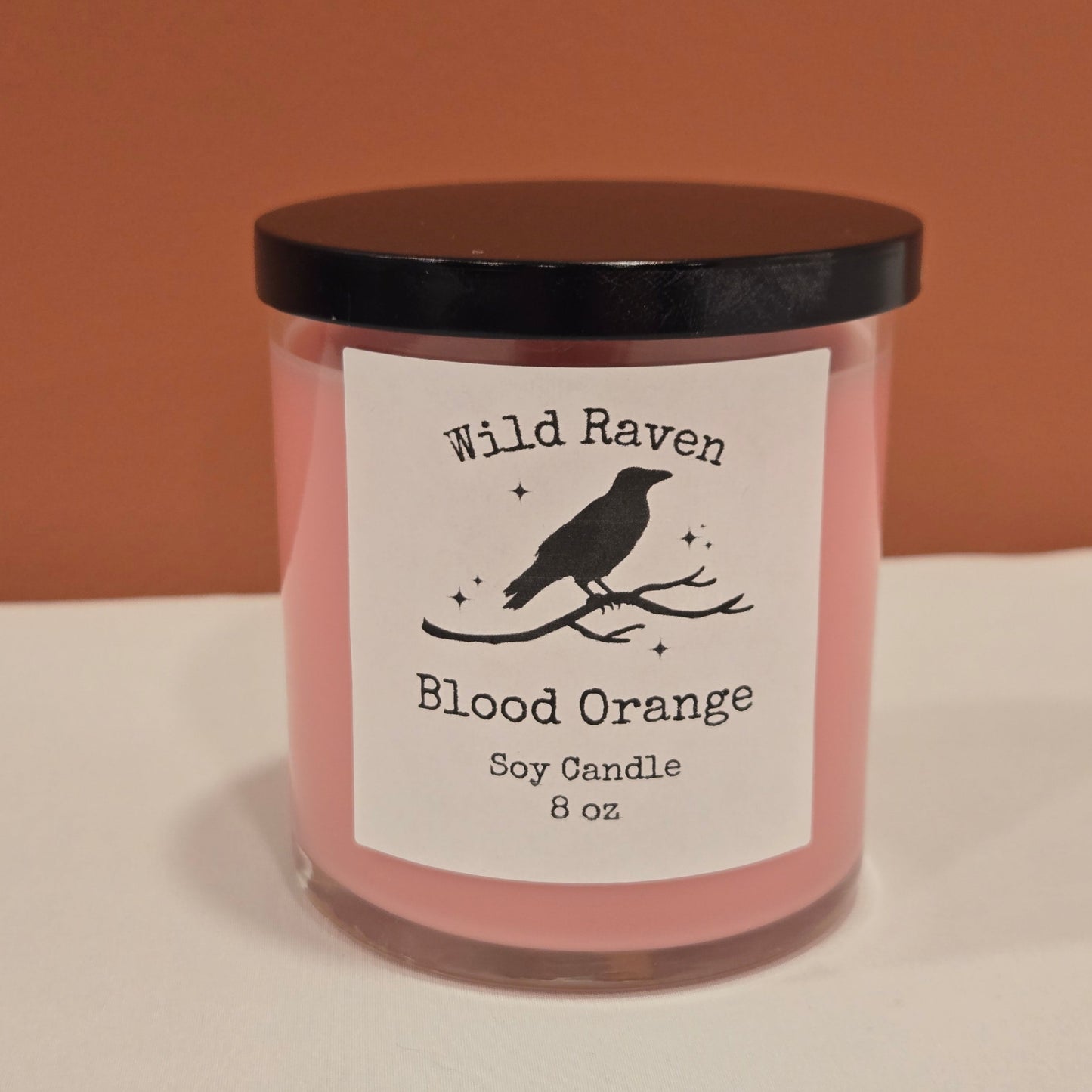 A pink soy candle in a clear glass jar with a black lid and a white label reading 'Wild Raven Blood Orange Soy Candle 8 oz' with a black bird silhouette, shown against a split brown and white background.