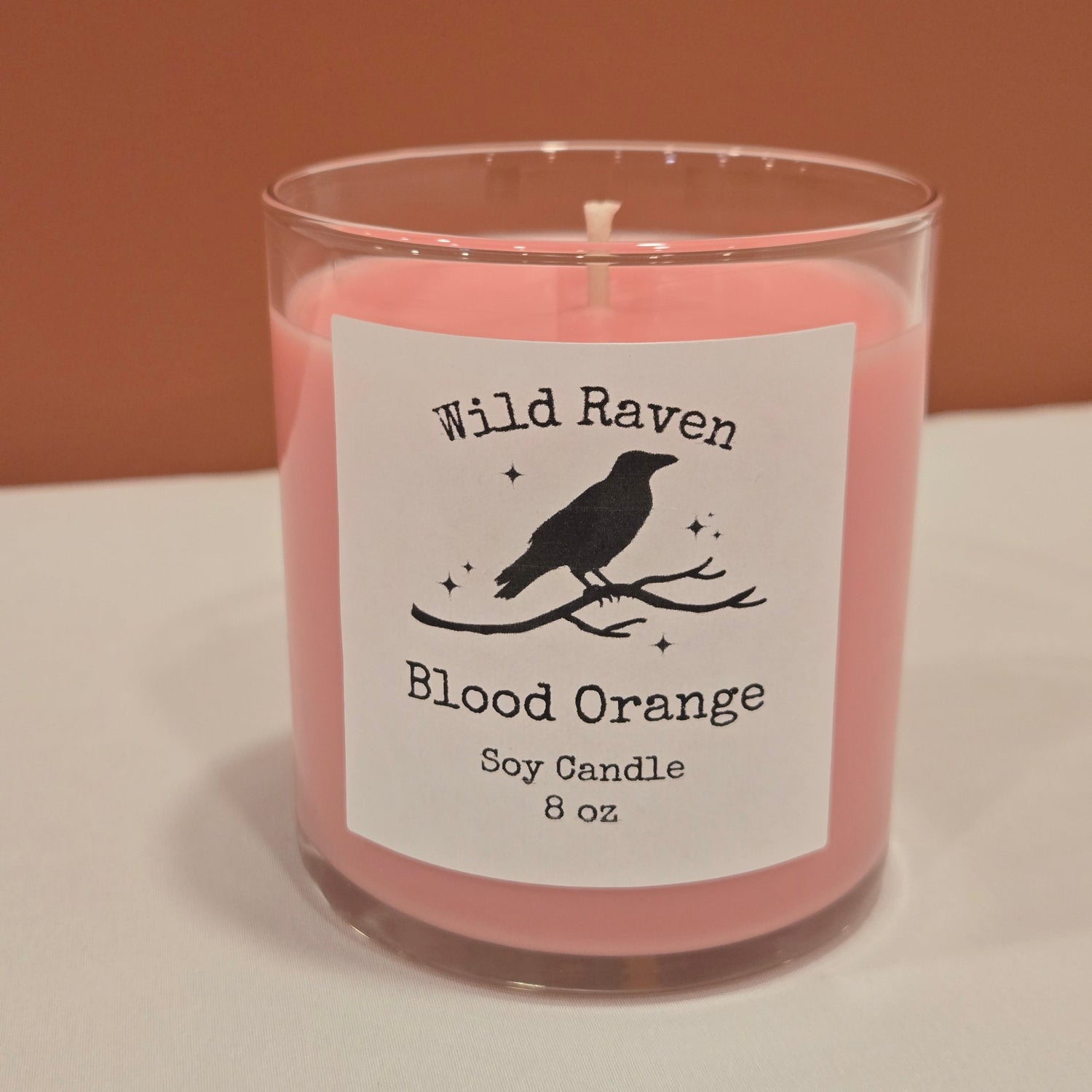 A slightly pink soy candle in a clear glass jar with a white label reading 'Wild Raven Blood Orange Soy Candle 8 oz' and a black bird silhouette, photographed against a split tan and white background.