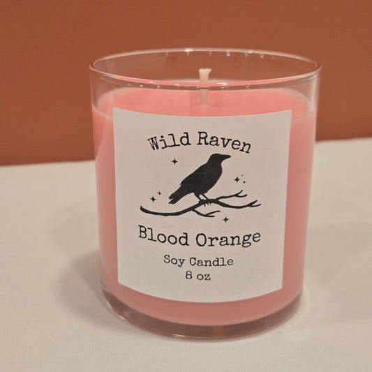 A slightly pink soy candle in a clear glass jar with a white label reading 'Wild Raven Blood Orange Soy Candle 8 oz' and a black bird silhouette, photographed against a split tan and white background.