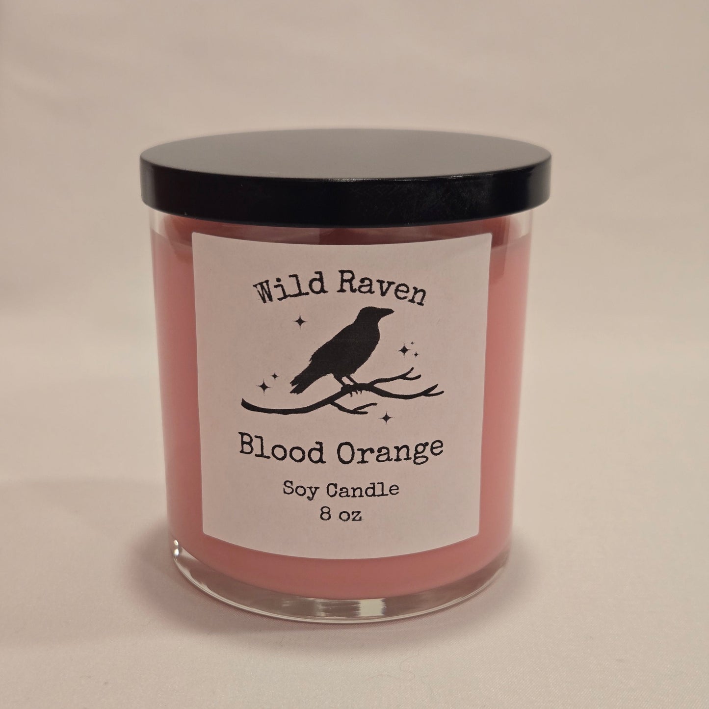 A 8oz soy candle in a clear glass jar with pink wax and a black lid, featuring a white label with a black bird silhouette and the text 'Wild Raven Blood Orange'.