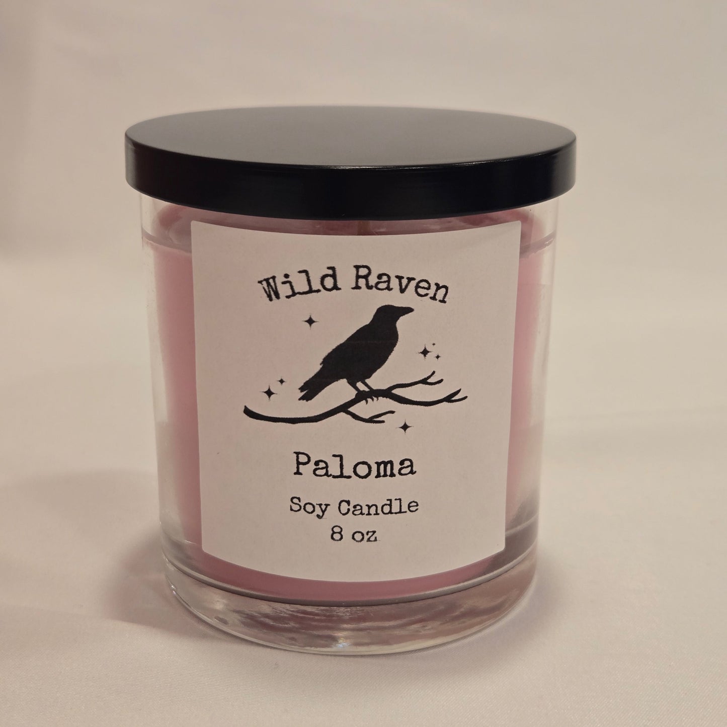 A 8 oz soy candle in a clear glass jar with a black lid and a white label featuring a black raven silhouette and the brand name Wild Raven.