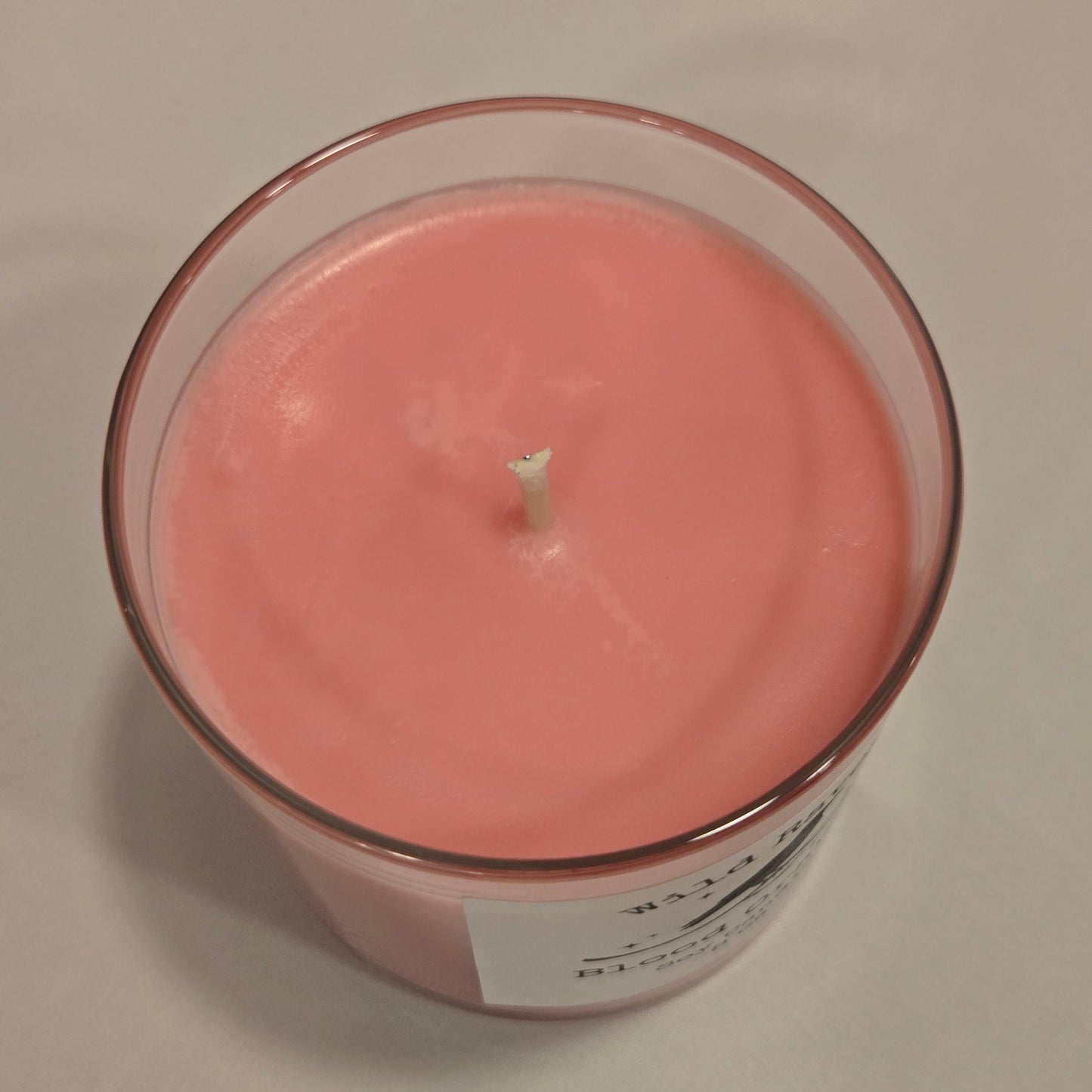 A  slightly pink and orange wax candle in a clear glass jar with a white label, viewed from directly above on a light beige background.