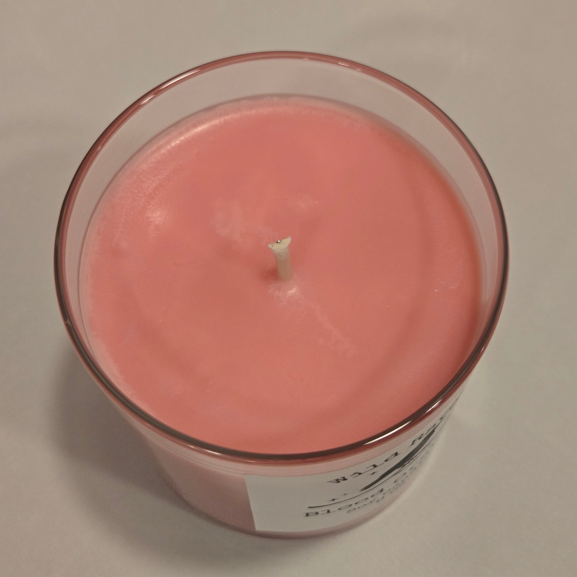 A  slightly pink and orange wax candle in a clear glass jar with a white label, viewed from directly above on a light beige background.